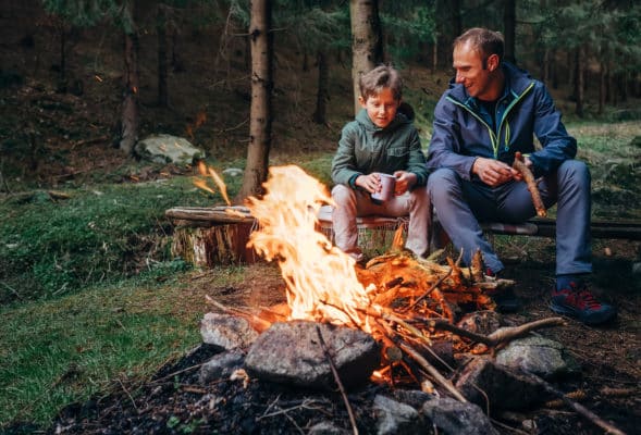 Father and son sit near a campfire in the woods in their permethrin-treated clothes.